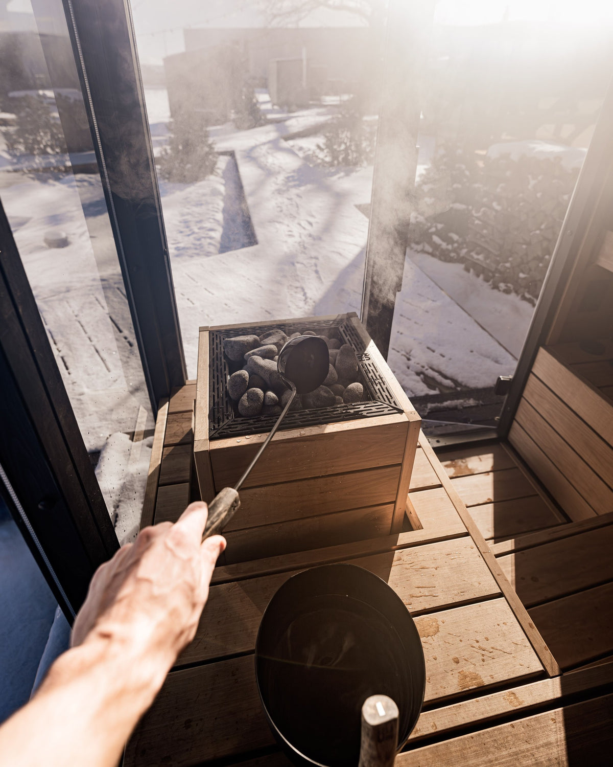 Pouring water over hot sauna stones, steam rising, with a snowy garden outside the glass